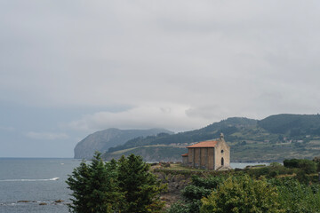 Alte Kapelle mit rotem Dach an der Atlantikk&uuml;ste im Baskenland, Nordspanien, umgeben von gr&uuml;ner Vegetation, Blick auf das Meer und bewaldete H&uuml;gel, bew&ouml;lkter Himmel, ruhige Stimmung.