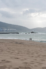 Zwei Personen stehen am Rand eines sandigen Strandes vor einer rauen See, im Hintergrund sind H&uuml;gel, Industrieanlagen und ein bew&ouml;lkter Himmel mit diffusem Licht zu sehen. Die Atmosph&auml;re wirkt ruhig u