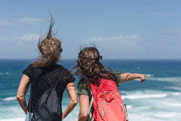 Zwei Frauen mit Rucks&auml;cken stehen an der windigen Atlantikk&uuml;ste im Baskenland, Nordspanien, blicken aufs Meer, eine zeigt nach vorne, Haare werden vom Wind verweht, sommerliche Outdoor-Stimmung.