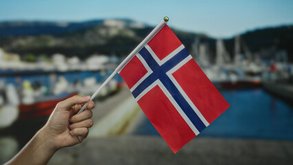 Hand holding norwegian flag against seaside port with blurred boats outdoors on a sunny day,...