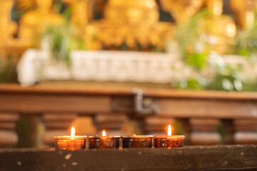 Mehrere brennende Teelichter stehen in einer Reihe auf einer dunklen Holzbank in einer Kirche. Der Hintergrund ist unscharf, golden und wirkt festlich, vermutlich ein Altar mit Dekorationen. Die Szene