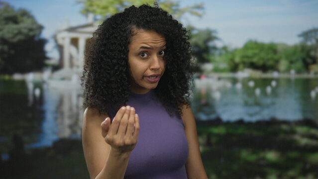 Young latina woman beckons with hand in green park by a pond near trees and walkway; welcoming invitation.