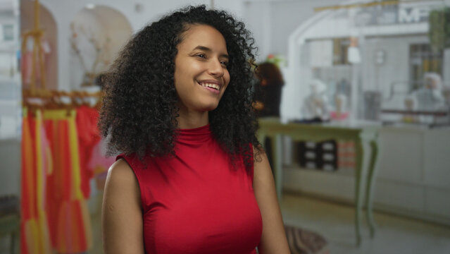 Hispanic woman in red top smiling inside a clothing store with vibrant dresses hanging in the background, suggesting a lively retail environment.