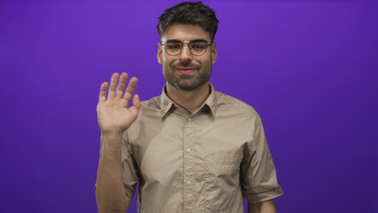 Hispanic man wearing beige shirt and round glasses waves hand in purple studio setting; friendly welcome.