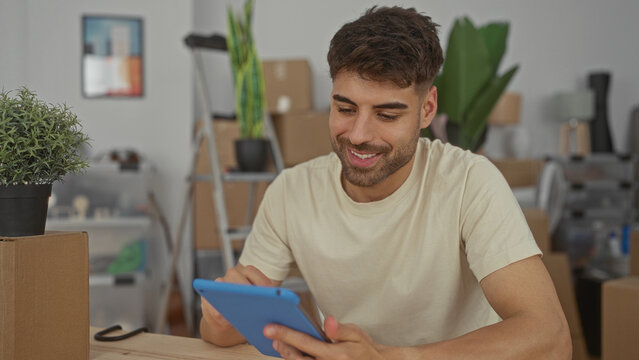 Man taps tablet screen with finger at wooden desk in building with moving boxes and potted plants; settling contentment.
