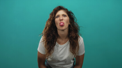 Fototapeta premium Woman holding both hands by her face and sticking out her tongue in studio teal backdrop; playful silliness joy.