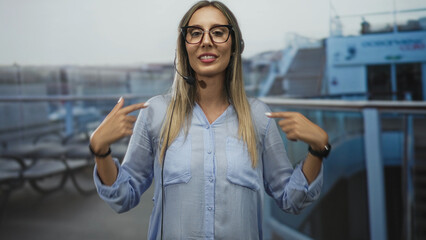 Woman call center operator wearing headset pointing fingers to chest on cruise ship deck with...