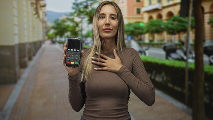 Woman young hispanic blonde holds portable card reader in city street with right hand raised showing device and left hand on chest, eyes closed; gratitude trust.