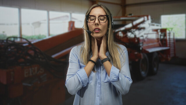 Young blonde woman call center operator touches neck while wearing headset in industrial building; work stress.