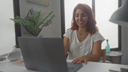 Woman waving hand at laptop in building desk workspace with headphones and potted plant visible while smiling during video call; remote work happiness.