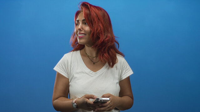 Woman smiling while typing on smartphone, hands and fingers visible, wearing white tee and necklaces in blue studio; happiness connection.