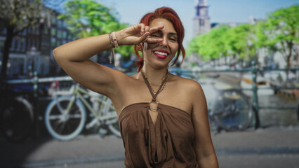 Woman smiling peace sign to eye on sunlit amsterdam street by canal and parked bicycles near a bridge; playful confidence.
