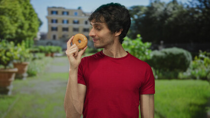 Fototapeta premium Young man smiles while holding a glazed donut near mouth at building entrance on a sunny day; happiness.