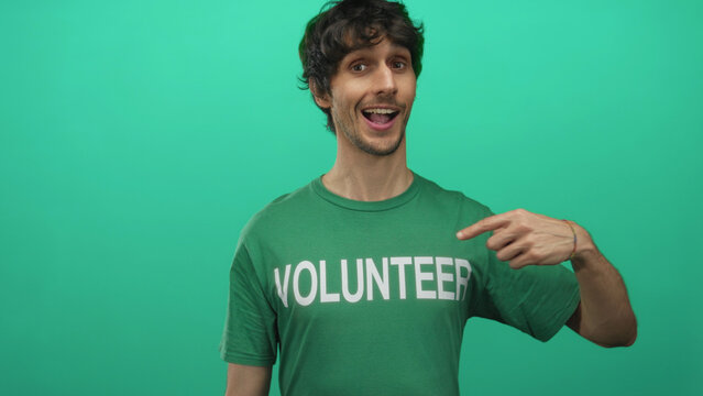 Young man smiling and pointing at volunteer shirt in studio while giving thumbs up and wearing green tee; support.