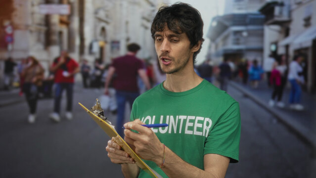 Man in green volunteer shirt holds clipboard and writes on form amid busy city street crowds; community service dedication support. - Powered by Adobe