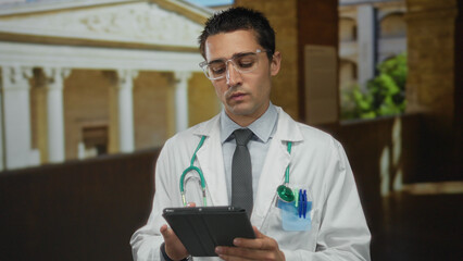 Young hispanic male doctor with stethoscope using tablet outdoors in old town, combining modern...