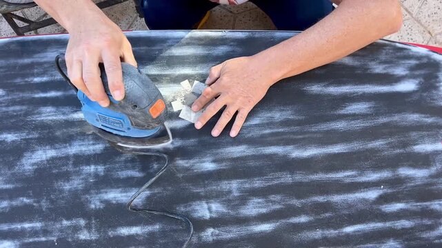 A man uses an electric sander to smooth the surface of a windsurf board outdoors, focusing carefully on detailed repair work