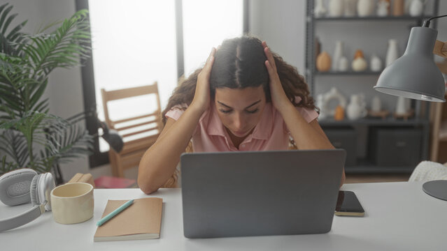 Woman holding head with hands over laptop at desk in studio, leaning forward near mug, notebook and smartphone; frustration focus.