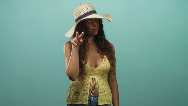 Woman pointing index finger upward in studio wearing straw sun hat and yellow knit vest while holding a small card; summer playful. - Powered by Adobe