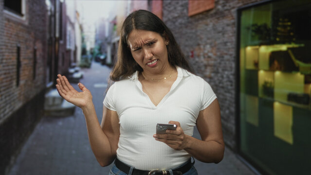 Young latina woman holding smartphone with puzzled frown, tapping screen and shrugging one hand in narrow city street; digital frustration.