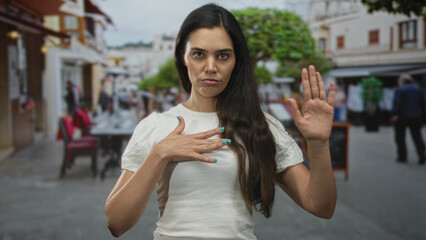 Woman young hispanic hand on chest and raised palm stop gesture in street cafe wearing white t shirt; oath solemn.