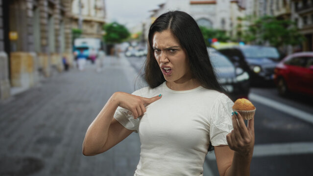 Woman holding muffin in one hand points finger to chest in a crowded street with buildings in view; confusion.