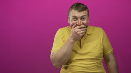 Man young caucasian with hand covering mouth wearing yellow polo in pink studio; stifled awkward amusement.