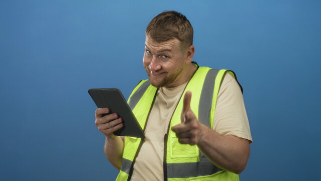 Man engineer wearing high visibility vest pointing finger at tablet in studio with blue backdrop; confidence.