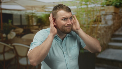 Man cups ear with hand on terrace near building entrance, young caucasian overweight with short beard and shirt sleeves, appearing strained; confusion listening.
