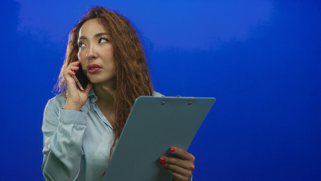 Woman holding clipboard and smartphone, reading notes while speaking on phone in blue studio; urgent concern.