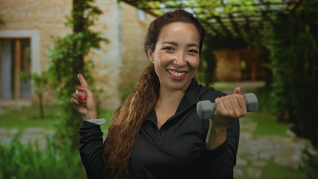 Woman points finger and holds dumbbell at house entrance outdoors while smiling and lifting weight; joy fitness motivation.