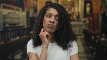 African american woman with hand on chin seated on wooden pew in church sanctuary; contemplation...