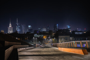 A nighttime view of Warsaw’s skyline captured from a modern footbridge, with shining skyscrapers and the Palace of Culture glowing in the distance.