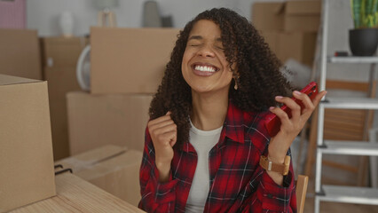 African american woman scrolling smartphone at office desk amid cardboard boxes and ladder; excitement.