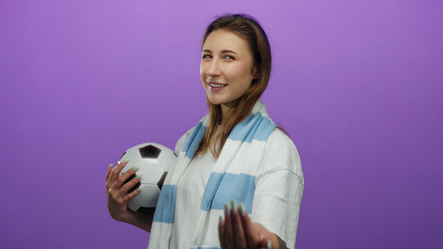 Woman holding soccer ball with inviting gesture and playful expression against vibrant purple wall, showcasing sports enthusiasm and casual style.