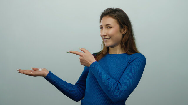 Young woman in blue sweater presents with open hand against a white isolated background, smiling confidently with a casual yet professional demeanor.
