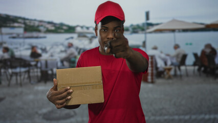 Man holds cardboard box beside restaurant tables on street by waterfront boats; service duty reliability.