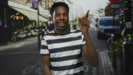 Man pointing finger upward with his right hand on a busy city street wearing a striped t shirt and smiling broadly; optimism inspiration motivation confidence.