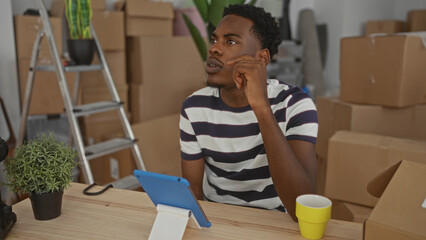 Young man holds tablet with finger on chin amid scattered cardboard boxes and potted plant on table in a building; contemplation.