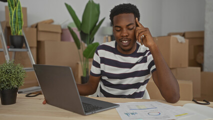 Man talking on phone in building while working on laptop among cardboard boxes; communication concentration.