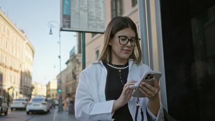 Woman young hispanic brunette tapping smartphone with both hands and visible fingers while standing by a building window on street in rome; focused.
