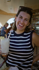 Woman holding a cold pineapple cocktail glass and sipping through a straw at a building bar table, smiling; casual summer relaxation.