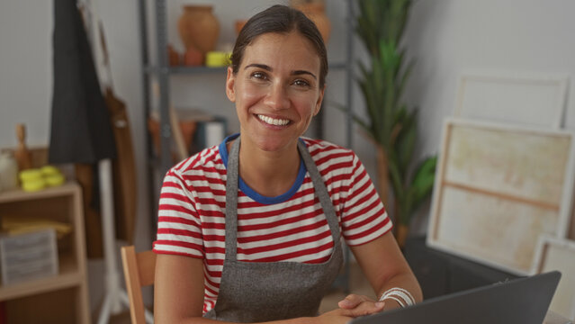 Woman smiling with hands clasped on table, wearing apron at a pottery worktable in studio surrounded by canvases and clay pots; creative joy.
