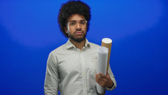 African american man with blueprints looks at watch, appearing focused in front of a solid blue wall while gesturing and holding plans.