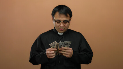 Priest holding american dollars excitedly against plain brown background