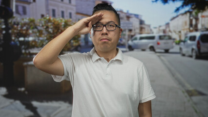Man in white polo salutes with right hand to forehead on a sunny street with parked vans and planters; duty.