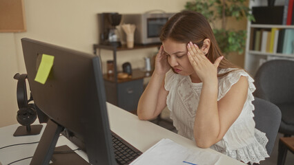 Obraz premium Young woman reading printed documents at desk by computer monitor and keyboard in building; stressed concentration.