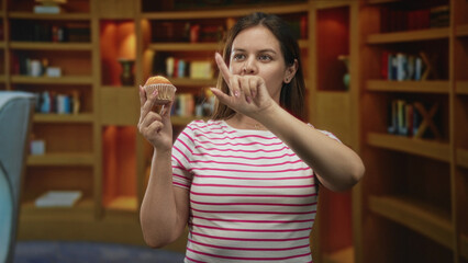 Woman in library holds a muffin in one hand and points finger to it while studying the pastry among bookshelves and an armchair, wearing a striped pink tshirt; doubt.
