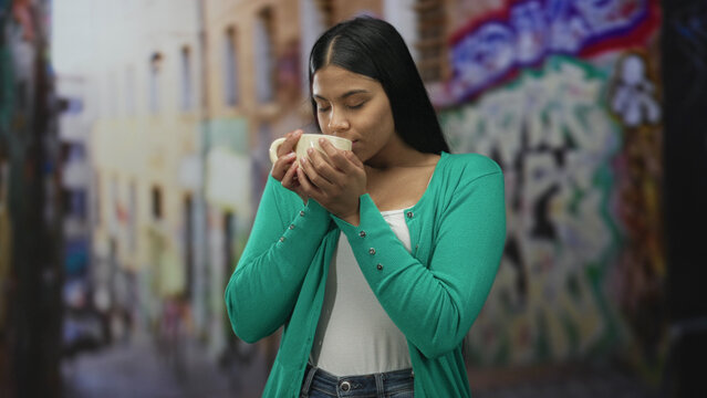 Young latin woman holding cup and looking up on urban street with colorful graffiti; calm reflection. - Powered by Adobe