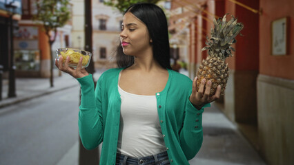Woman holding pineapple in one hand and bowl of chips in other while comparing on street; health...